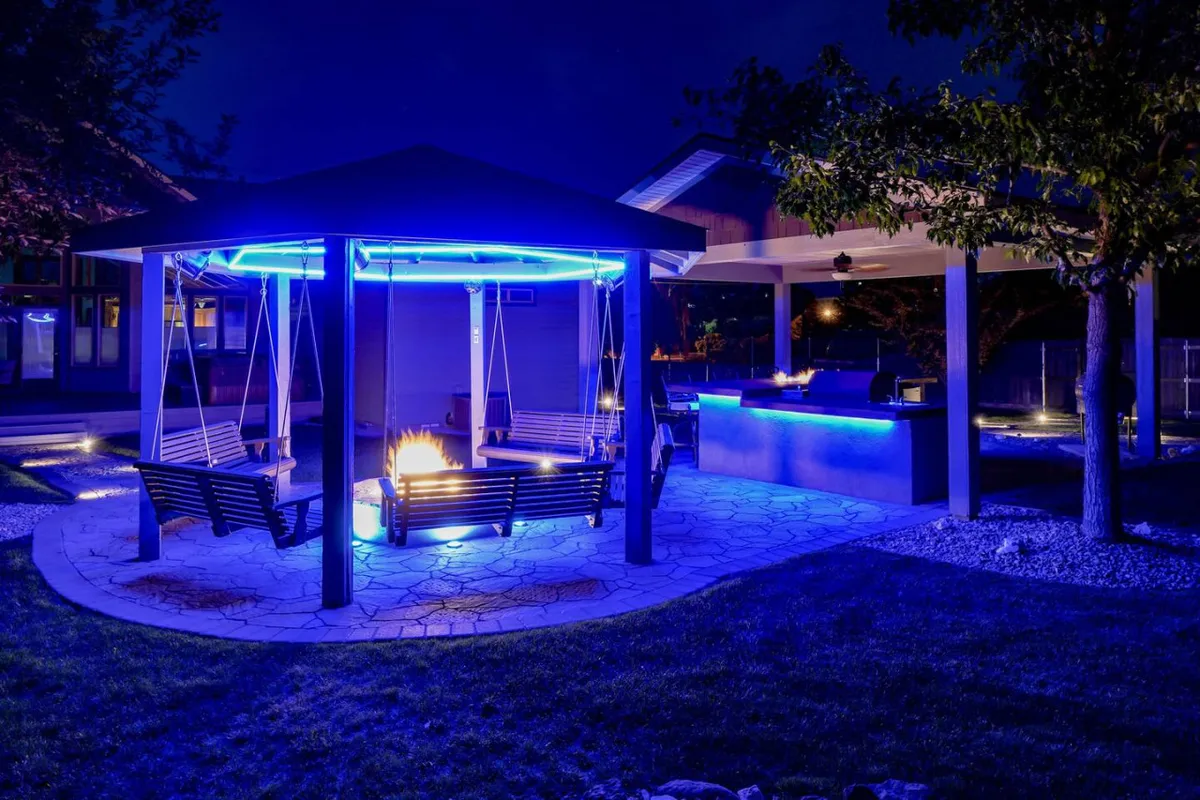 An inviting outdoor living area with an integrated swing set adorned with string lighting, a fire pit surrounded by comfortable seating, and an illuminated outdoor kitchen. The setting suggests evening gatherings, outdoor dining, and intimate conversations under the stars.