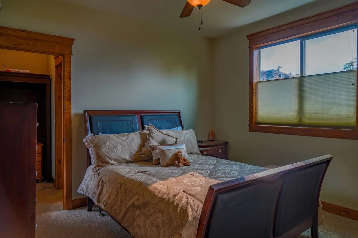 A calming bedroom scene with a bed adorned with decorative pillows set beside a window. Gentle lighting gives the room a cozy, serene vibe, perfect for quiet reading or restful sleep.