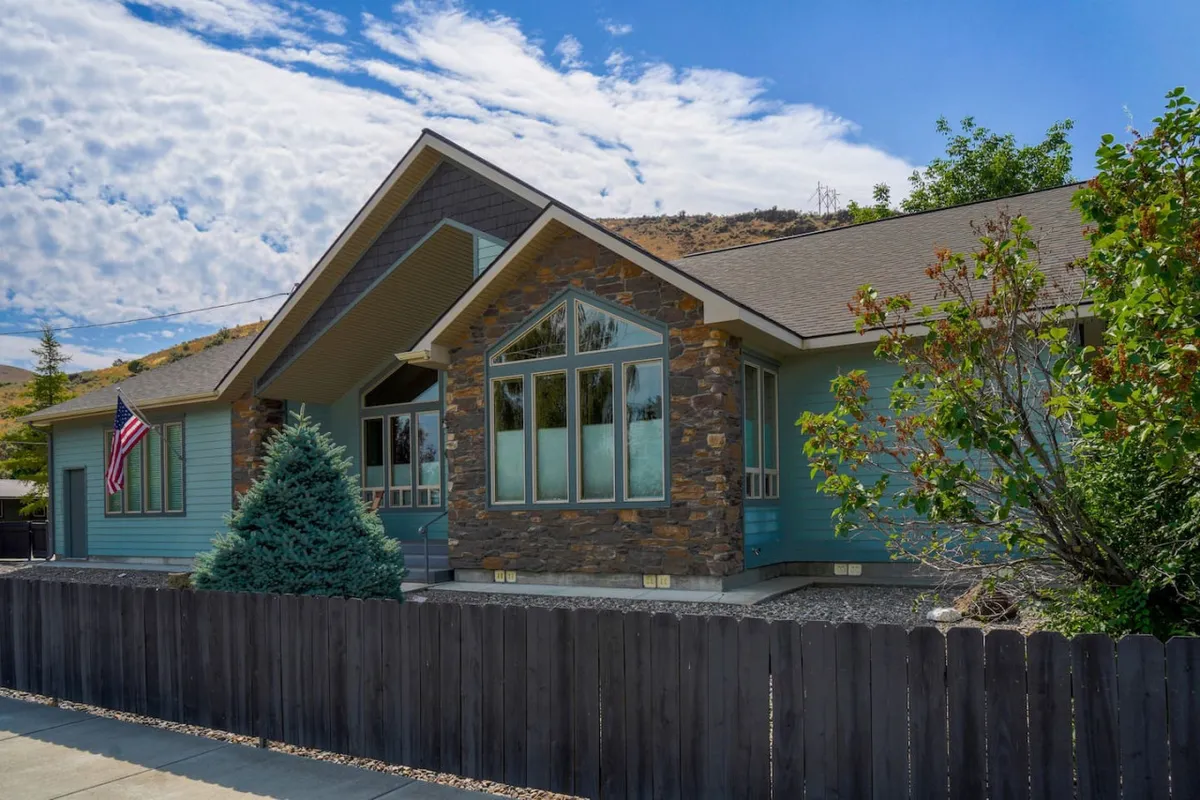 The exterior of Wunder House features a welcoming stone facade, large front windows, an American flag, and neatly trimmed shrubbery. The inviting yard and porch suggest spaces for relaxation and gatherings, with the mountain air visible in the background.