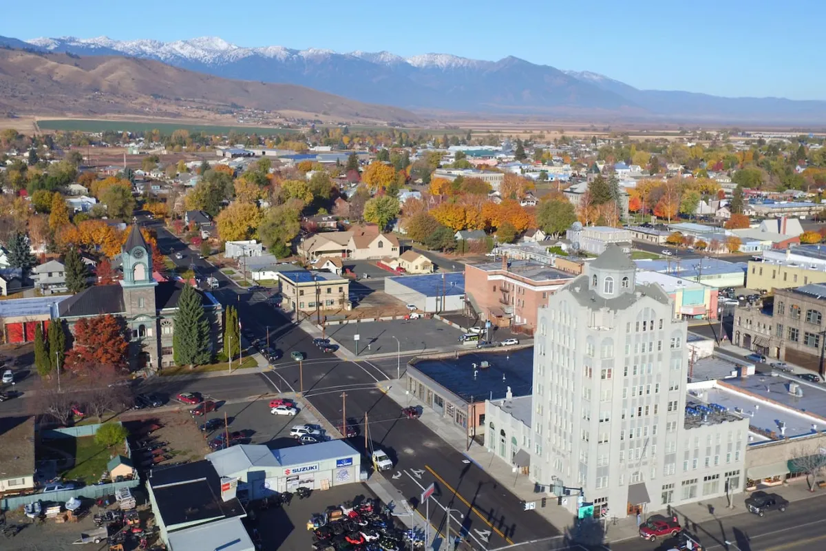 A scenic view of downtown Baker City with a prominent historic building, colorful autumn trees, and snow-capped mountains in the distance. The lively atmosphere hints at local shops, cafes, and a vibrant community environment.