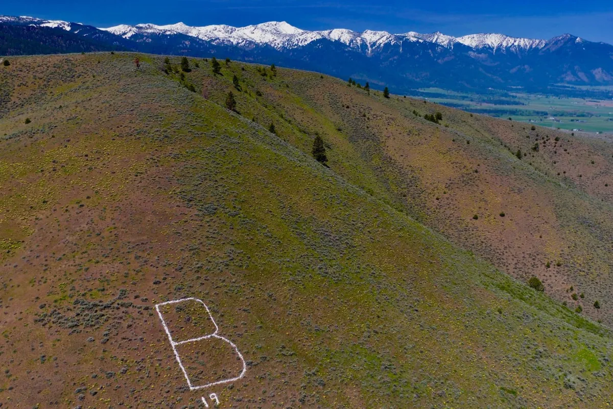 A sweeping view of snow-capped mountains rising above green hills, with the distinctive white letter 'B' visible on the mountainside. The landscape suggests the natural beauty and outdoor adventure that define the Baker City area and its proximity to Anthony Lakes.