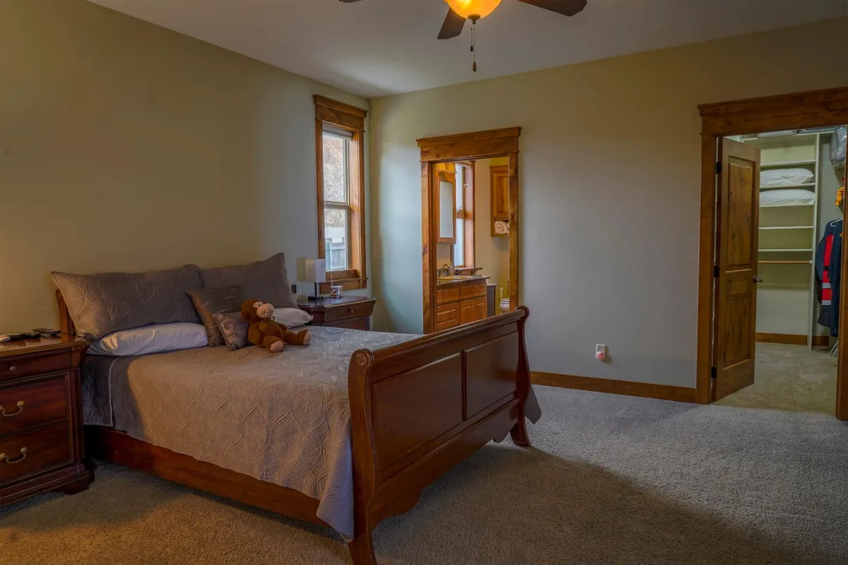 A peaceful bedroom featuring a neatly made bed, a nightstand, windows allowing natural light, and a doorway leading to another area. Soft colors and plush bedding create an inviting and restful environment ideal for unwinding.