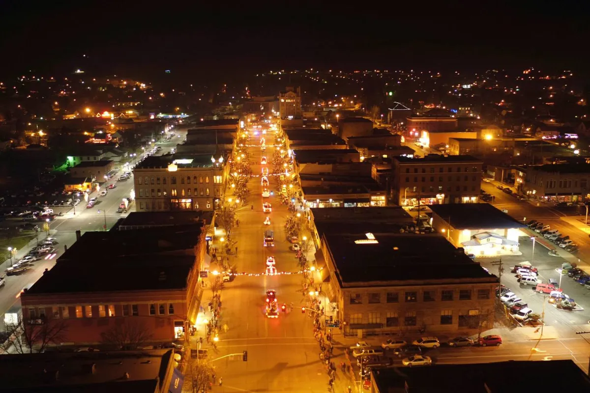 A festive street scene in downtown Baker City, featuring a street lined with glowing lights, vehicles participating in a parade, and historic brick buildings housing various shops and restaurants. The atmosphere is lively and celebratory, capturing the spirit of community events and local exploration.