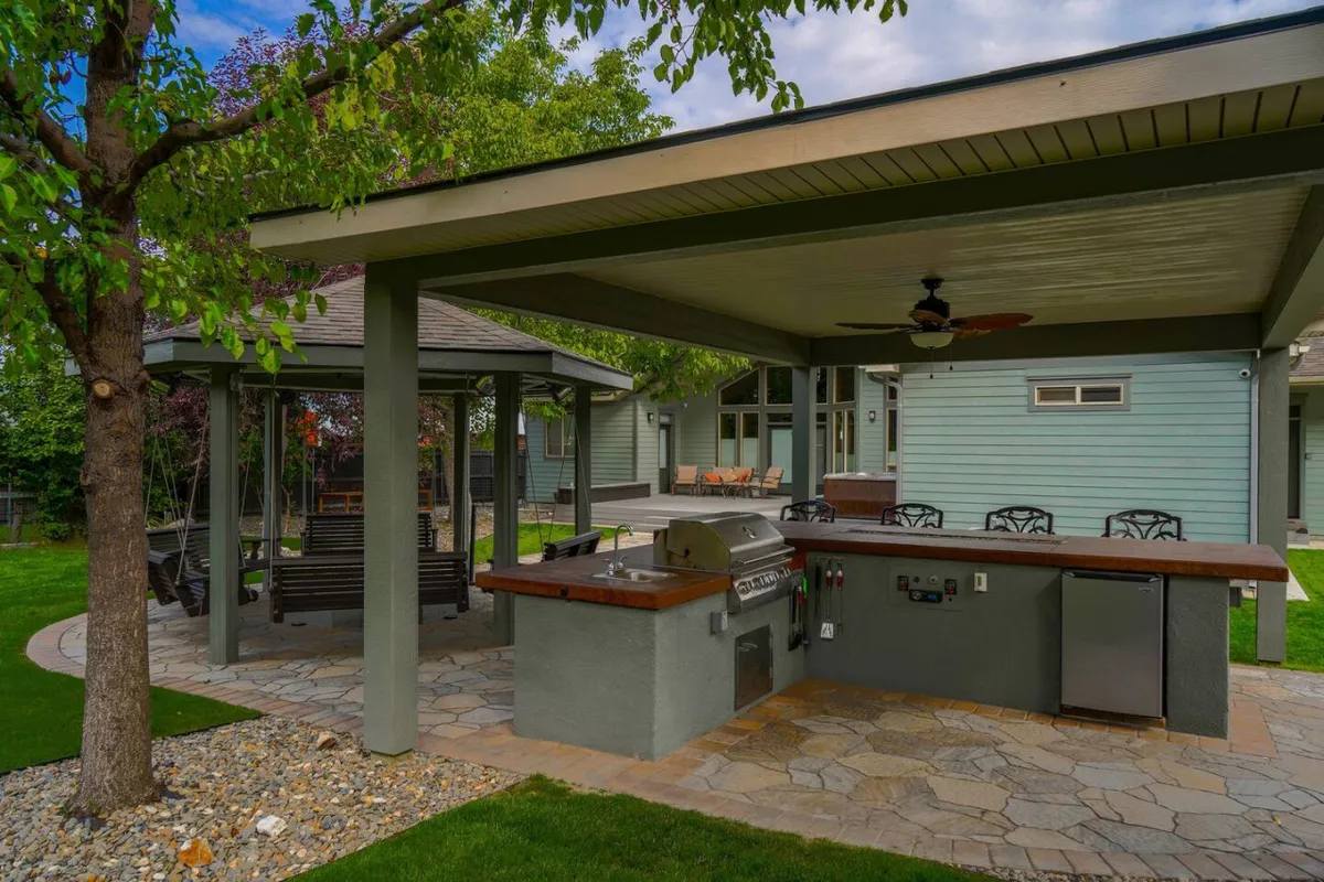 The photo focuses on a well-appointed outdoor kitchen area beneath a canopy of trees, complete with a built-in grill and BBQ setup. A swing adds a touch of playful relaxation to the scene, while the kitchen area is clearly designed for hosting and socializing.