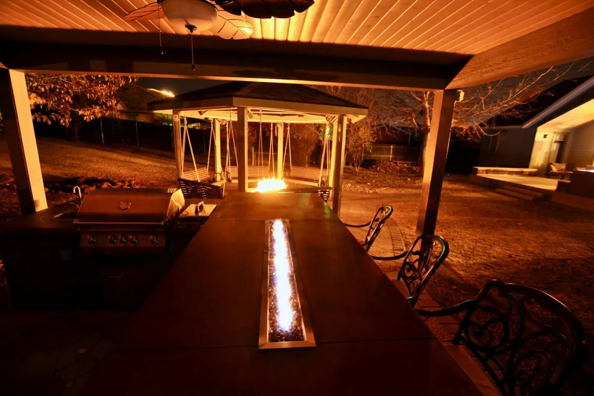 A warm and inviting outdoor kitchen scene with a grill, fire feature, swing seating, and a dining table. The setting is perfect for barbecues and gatherings, evoking memorable evenings with friends or family around good food and cozy firelight.