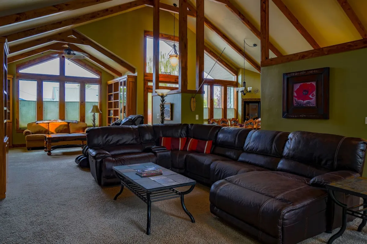 A cozy living area with a large leather sectional sofa, a wooden coffee table, large windows allowing generous natural light, and artistic wall decor. The arrangement encourages conversation and relaxation, embodying the warmth and comfort of communal living.