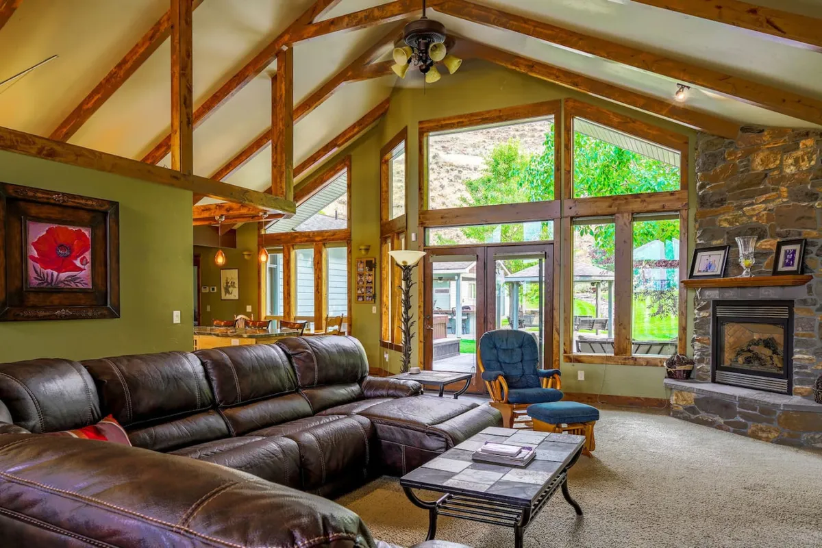 The image showcases a spacious, sunlit living room with a large leather couch facing a cozy stone fireplace. Wooden beams span the ceiling, adding rustic charm, while expansive windows fill the space with natural light. The room feels inviting, perfect for relaxation or gathering with family and friends.